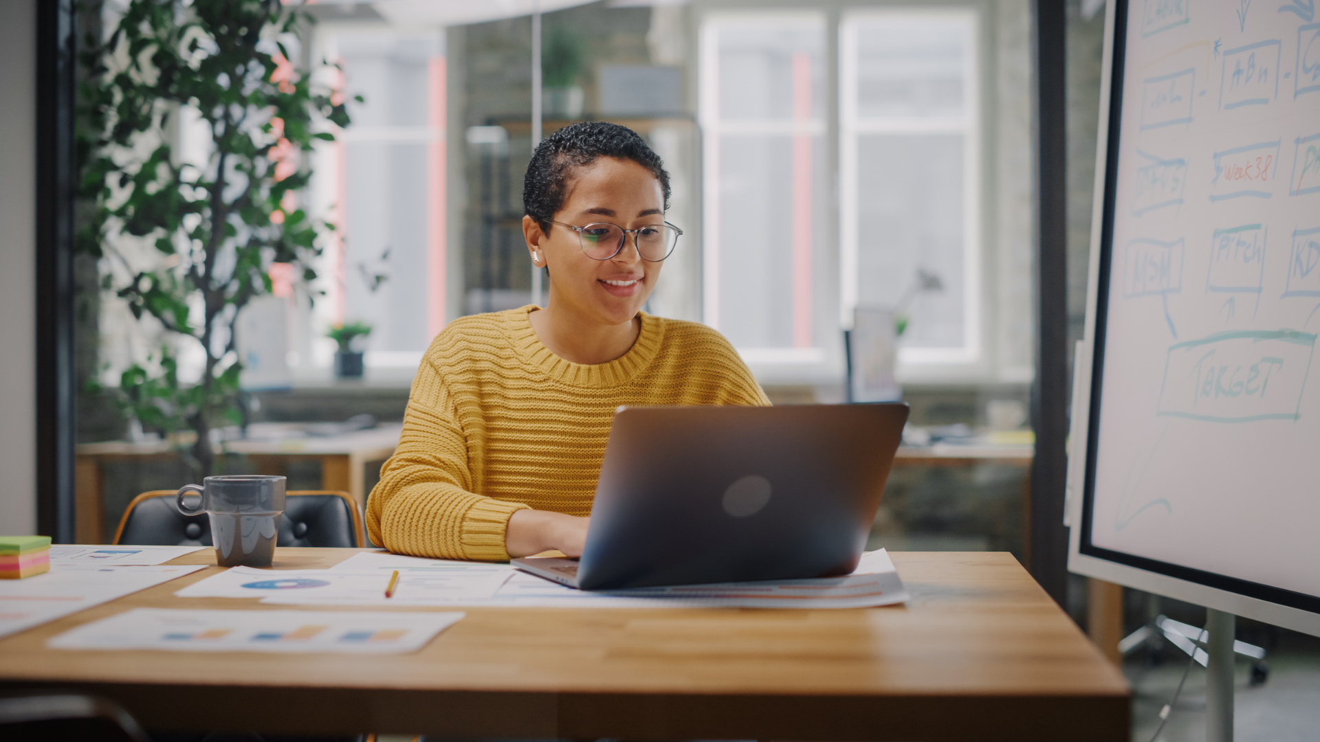 A smiling woman at a table with charts and diagrams, working on a laptop.